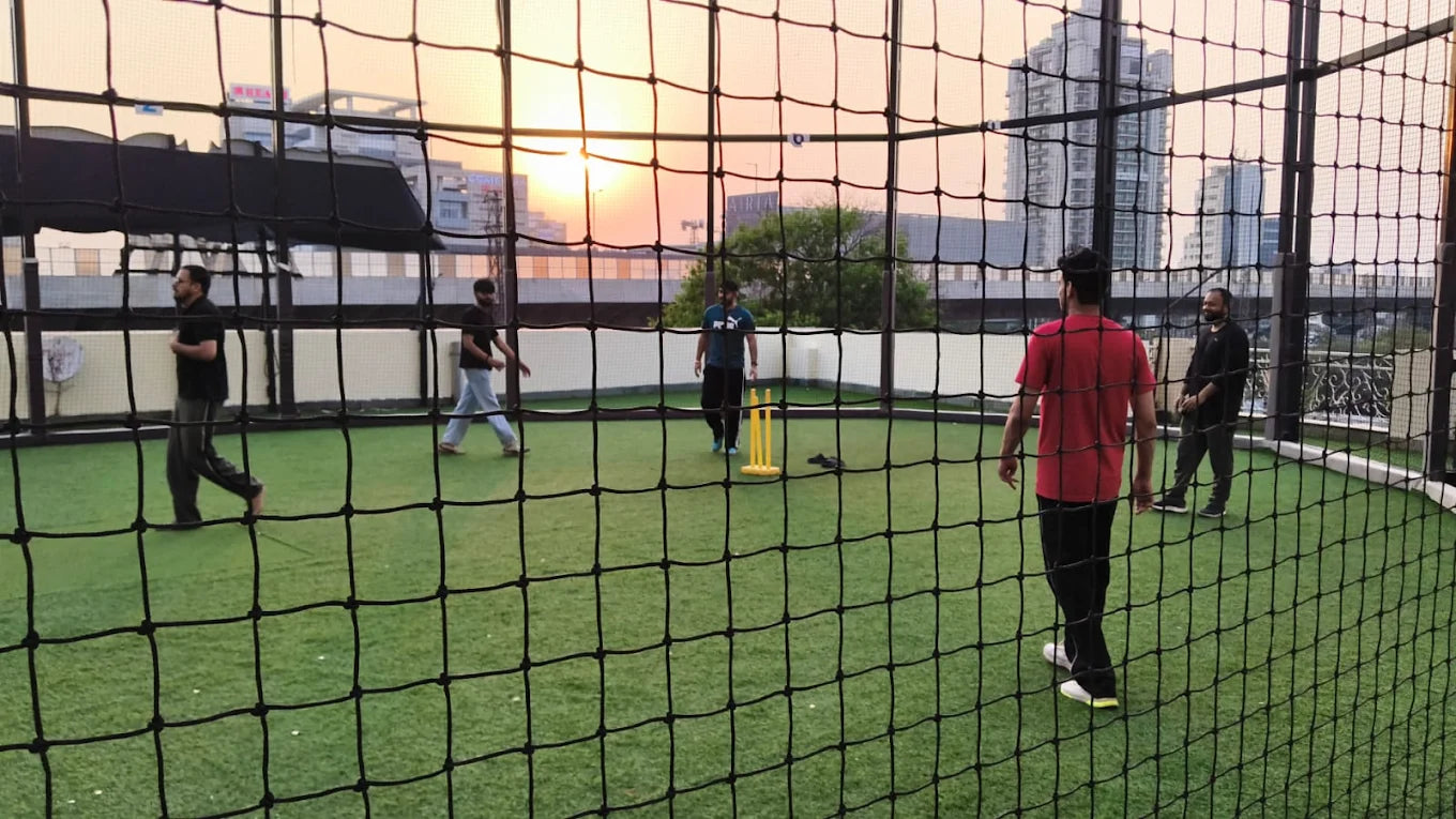 Adults play cricket on a rooftop field at sunset, with city buildings in the background.