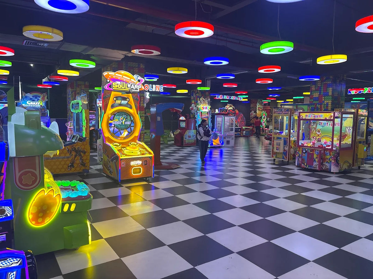 An arcade with colorful lights and checkered floors, featuring various game machines and a few young people.
