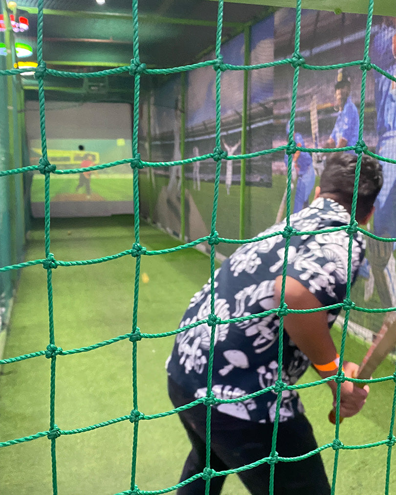 A young person plays cricket in an indoor batting cage, viewed through a green net.