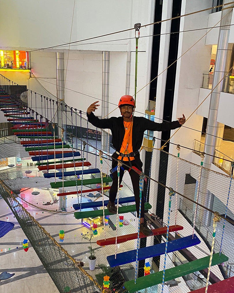 A young person in a helmet and harness navigates a rope bridge obstacle course in an indoor adventure park.
