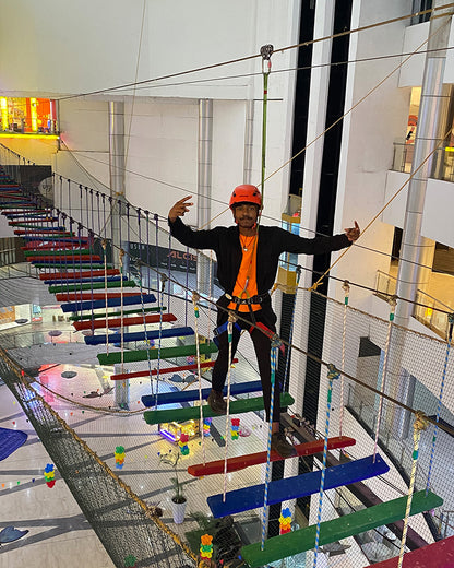 A young person in a helmet and harness navigates a rope bridge obstacle course in an indoor adventure park.
