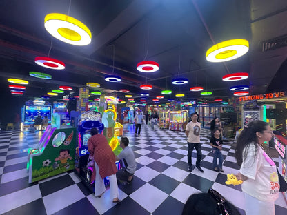 People play arcade games in a brightly lit entertainment center with a checkered floor and colorful hanging lights.