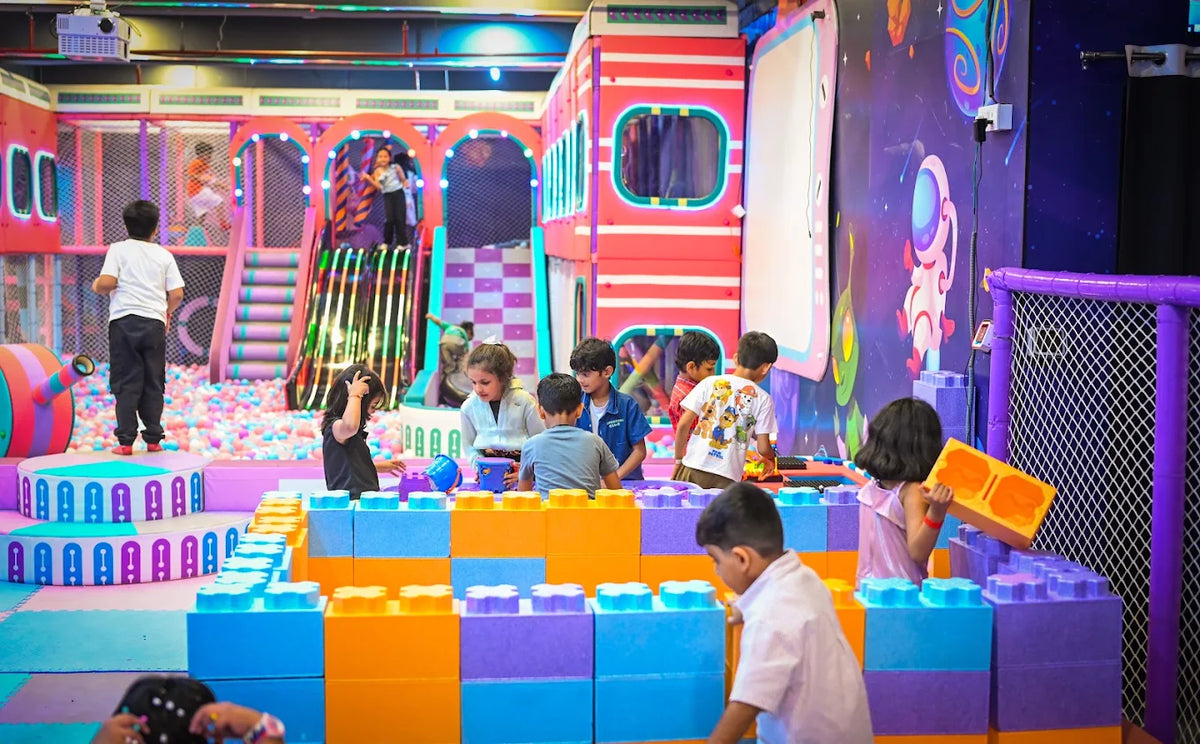 Children play with large building blocks in a colorful indoor playground with a ball pit and slides.