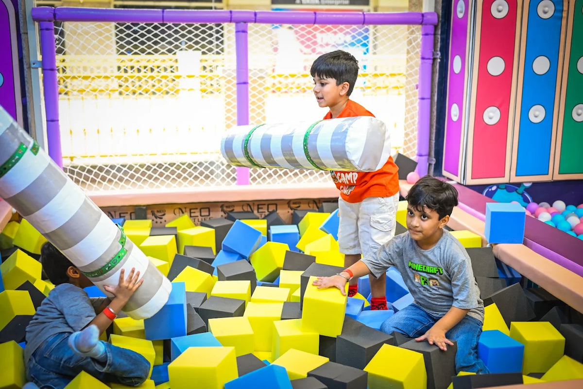 Two young boys play in a foam pit filled with colorful blocks, one holding a large cylindrical pillow.