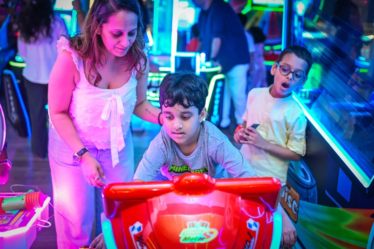 A woman watches two young boys play a racing arcade game in a brightly lit arcade.