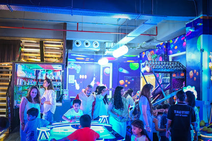 A group of adults and children enjoy arcade games in a brightly lit, neon-colored entertainment center.