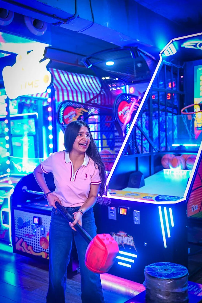 A young woman laughs while playing a carnival game in a brightly lit arcade. She holds a large mallet, ready to strike a target.
