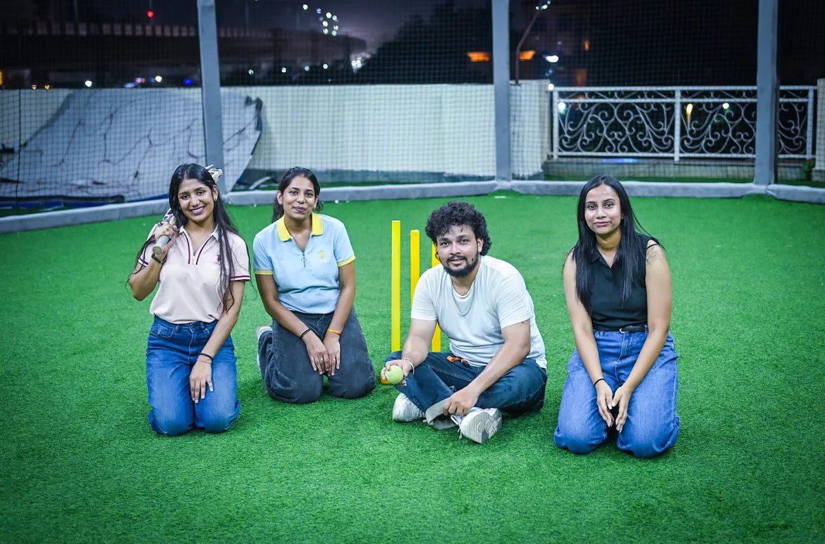 Four young adults kneel on a green turf field, two women holding a cricket bat and ball, with yellow wickets behind them.