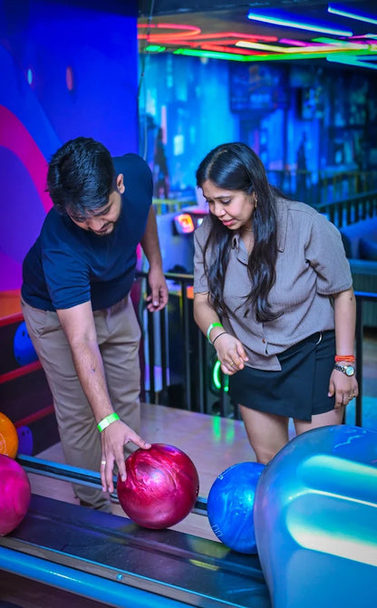 A young man and woman at a bowling alley, with the man reaching for a red bowling ball.