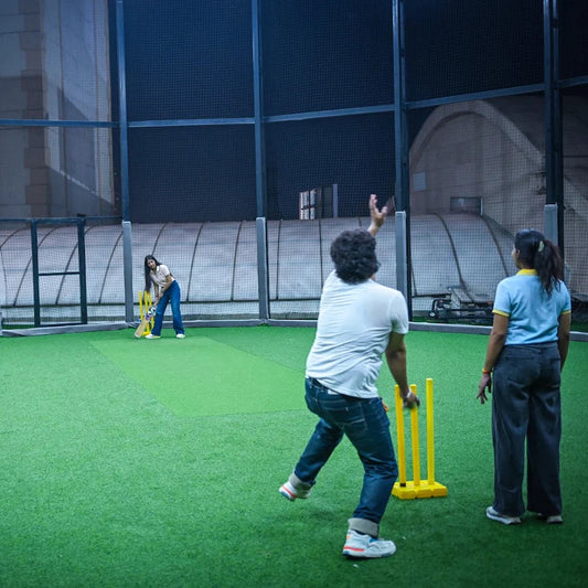 Adults play cricket on a green turf field indoors.