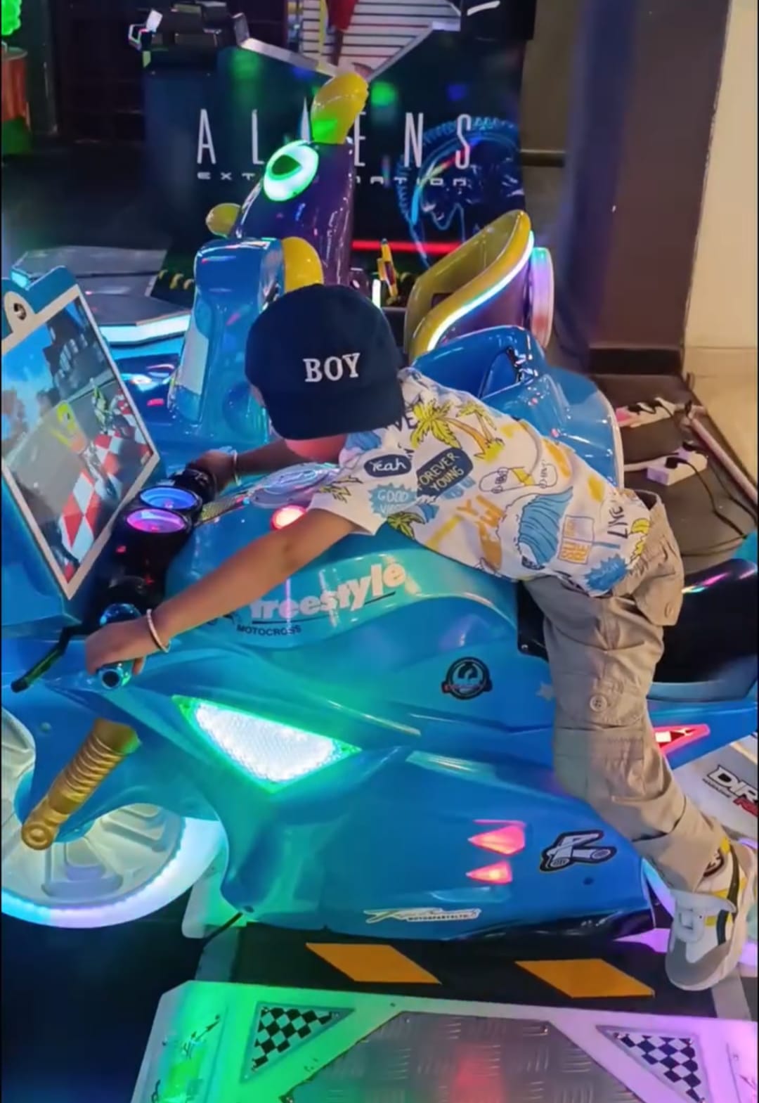 A young boy in a blue motorcycle arcade game. He wears a "BOY" hat and a patterned shirt.