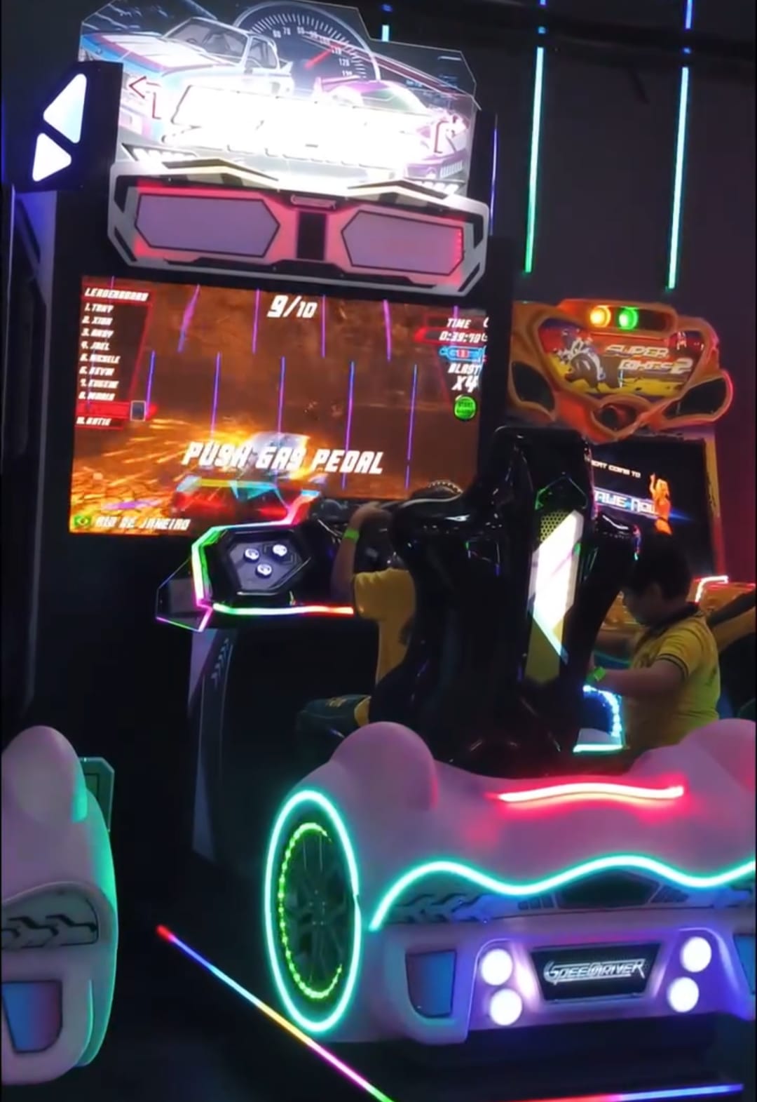 A child plays a racing arcade game in a dimly lit arcade, with neon lights illuminating the game's car-shaped seat.