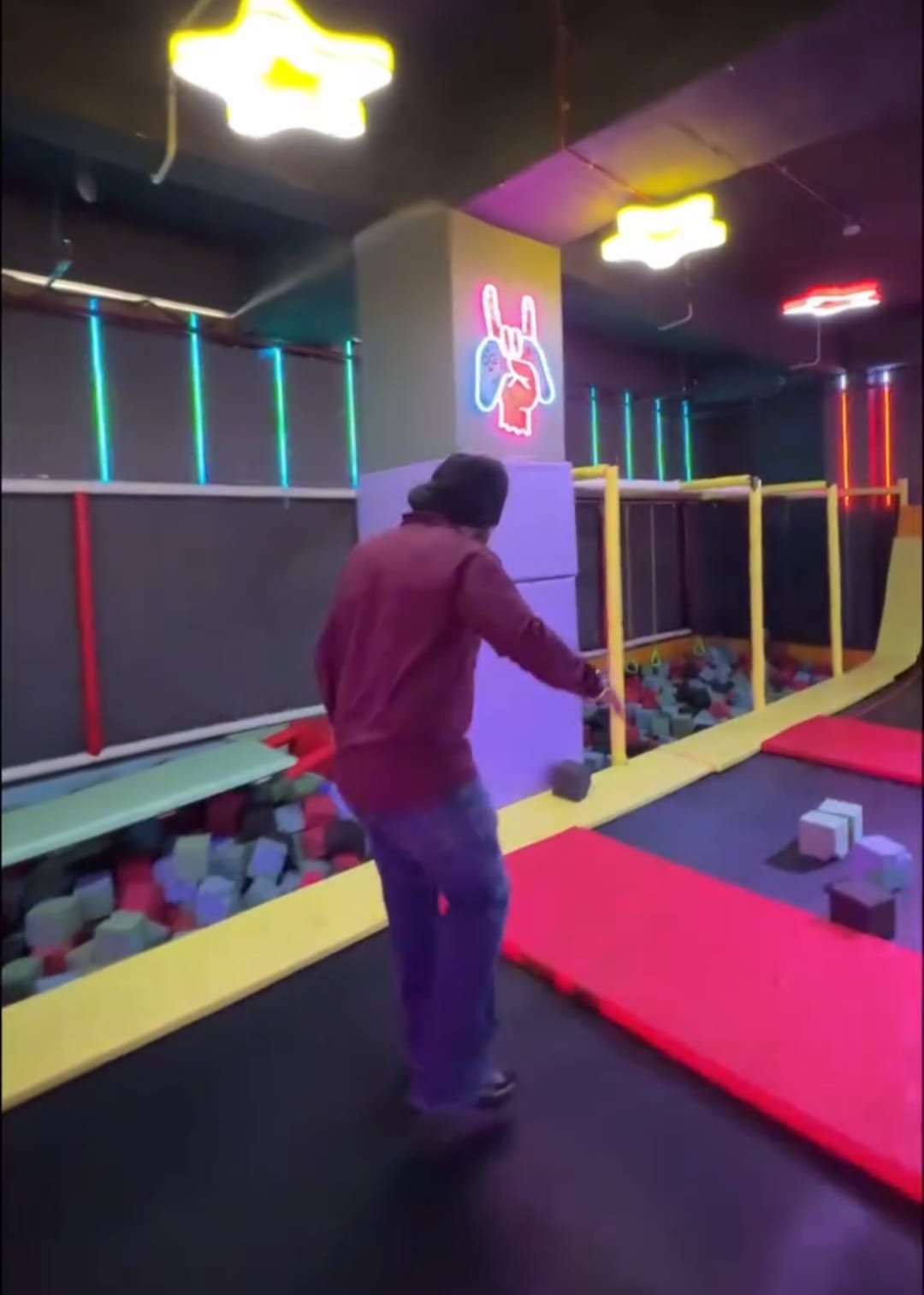 A person jumps on a trampoline in a colorful indoor play area with foam pits and neon lights.