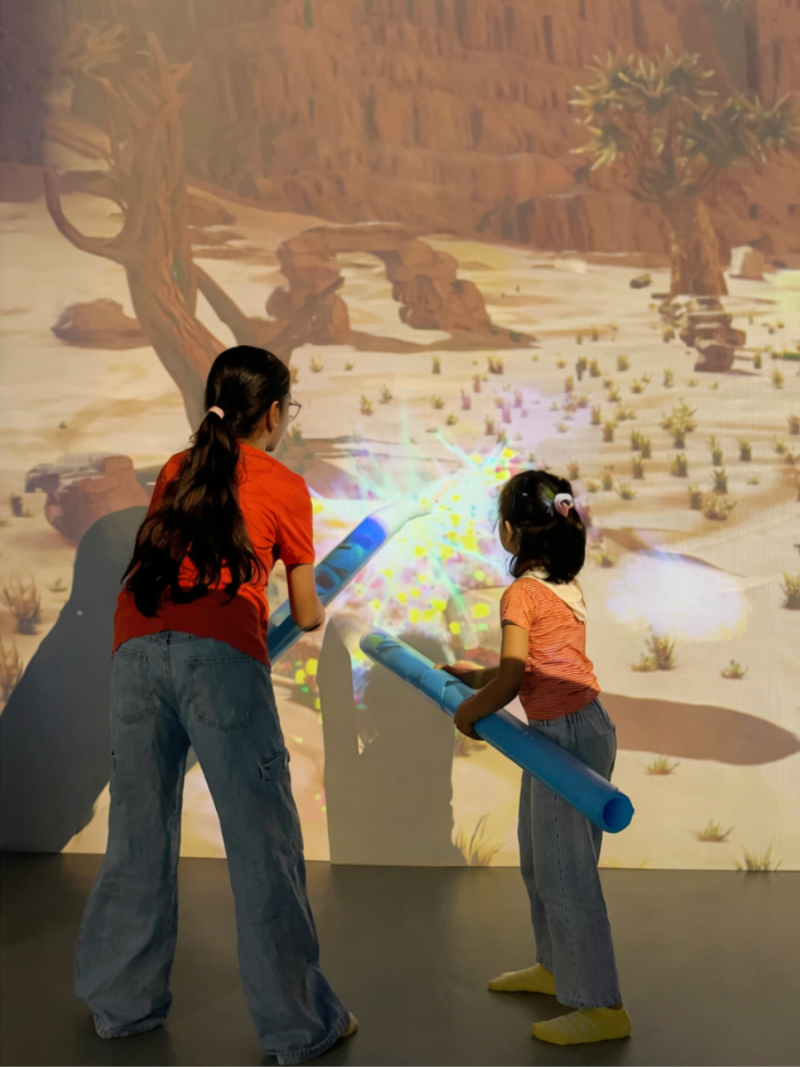 Two young girls play with light-up tubes in front of a projected desert landscape.