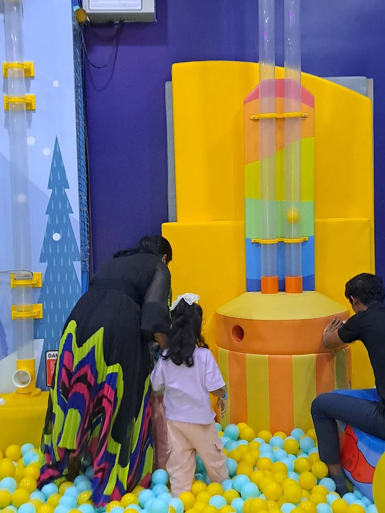 A woman and two children play in a ball pit with colorful tubes and structures.