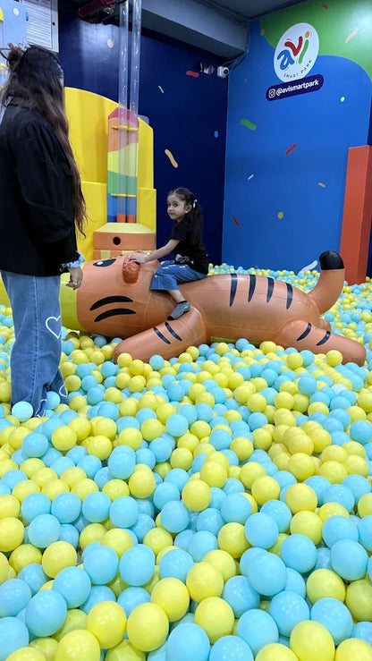 A young child sits on a tiger inflatable in a ball pit, with an adult standing nearby.