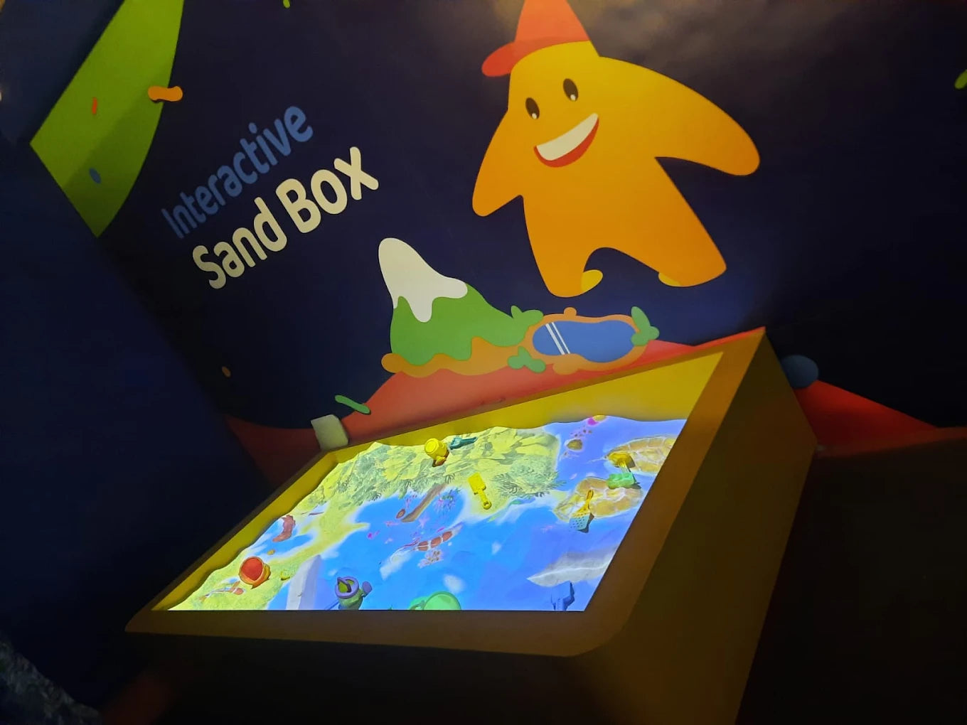 An interactive sand box with a projected landscape of water and land, featuring cartoon decorations for children.
