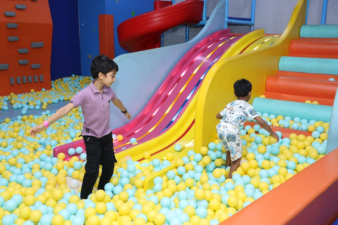 Two young boys play in a ball pit with a colorful slide in the background.