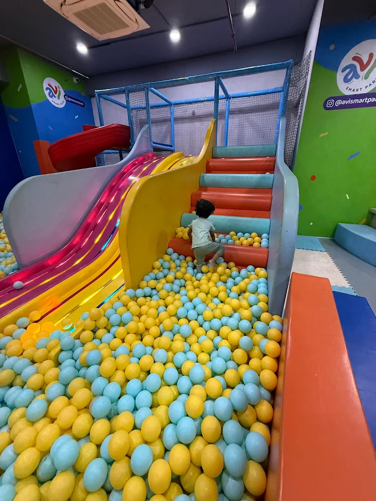 A young child climbs stairs in a ball pit with a colorful slide.
