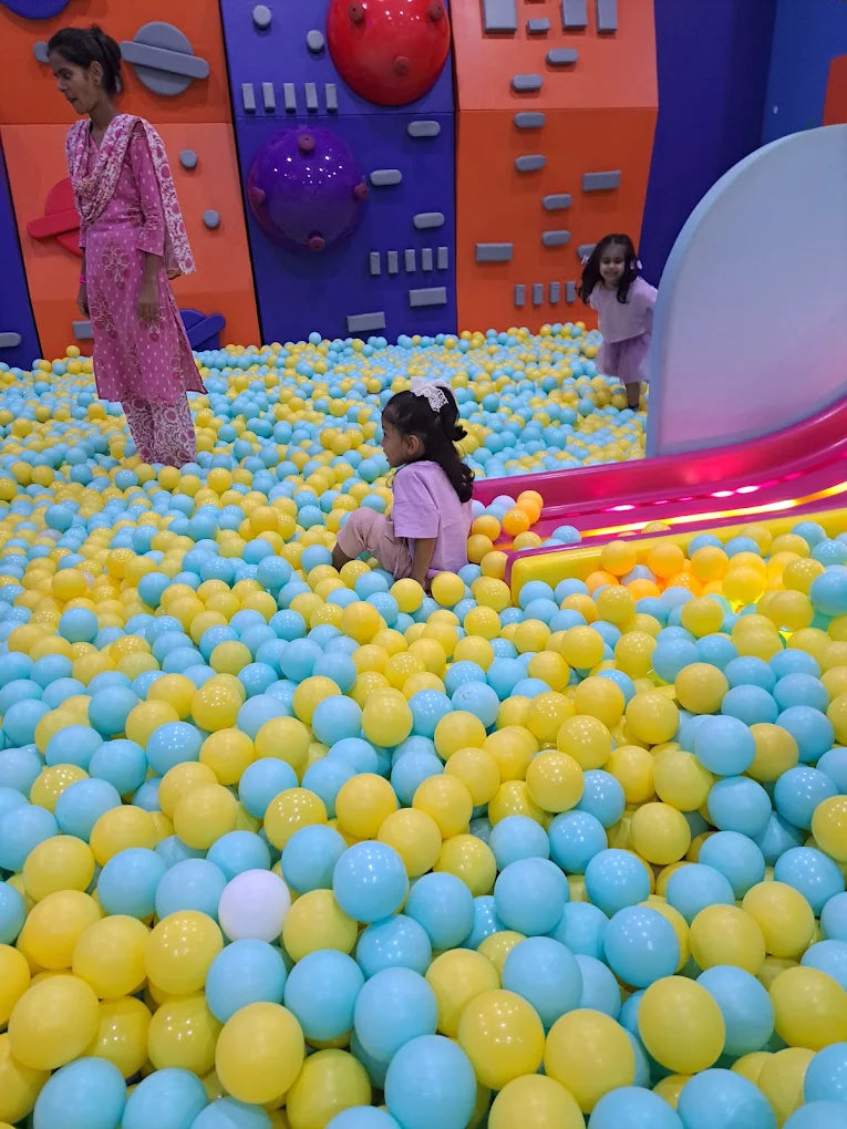 Children play in a ball pit filled with blue and yellow balls, with a woman standing nearby.
