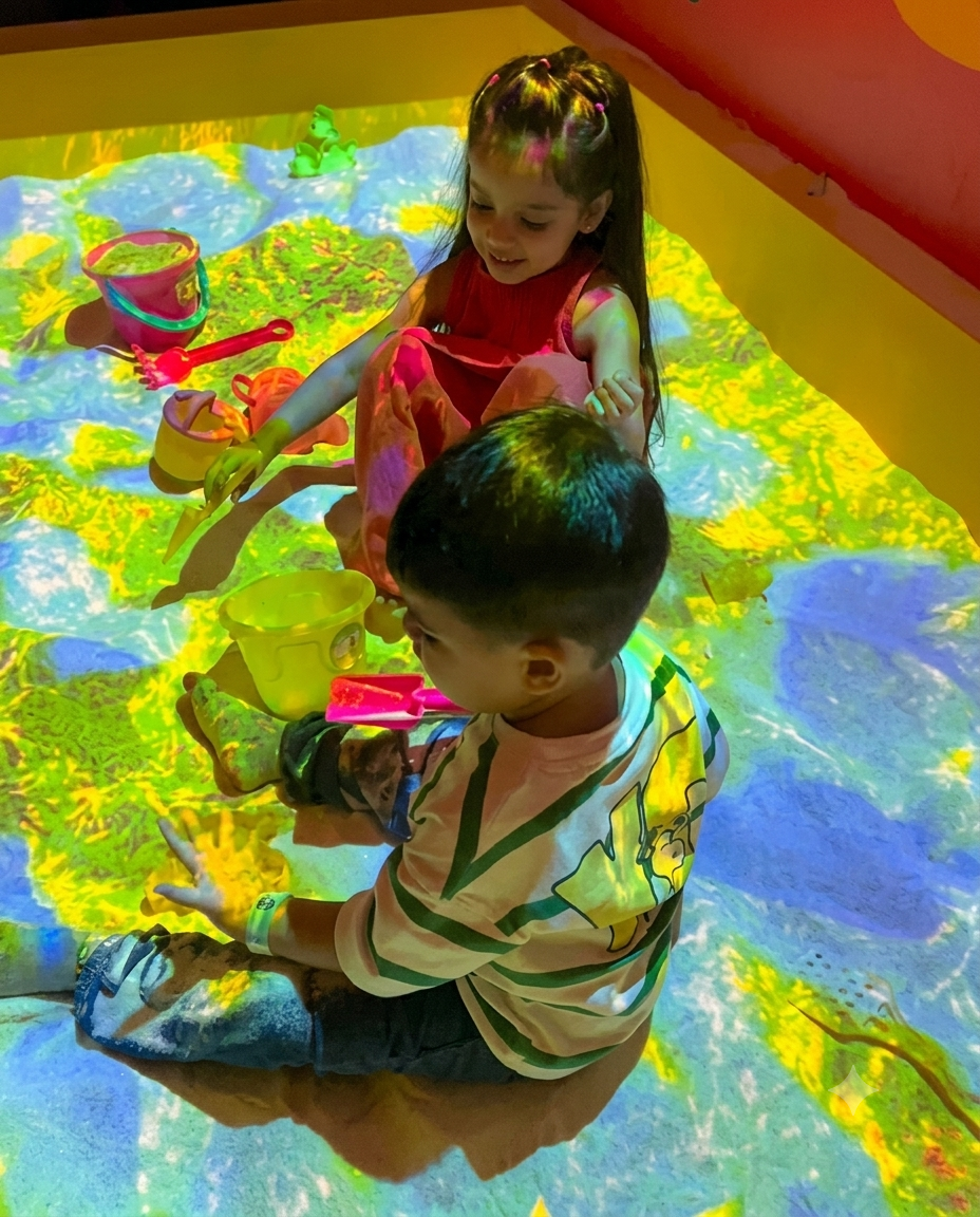 Two young children play with sand toys on a projected, colorful landscape.