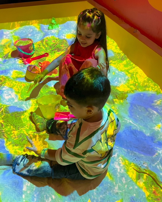 Two young children play with sand toys on a projected, colorful landscape.