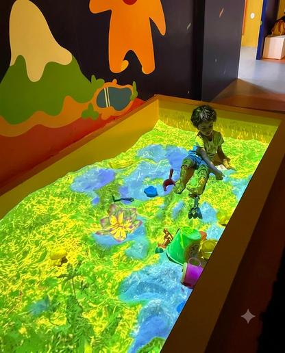 A young child plays in a sandbox with projected interactive graphics of a vibrant landscape.