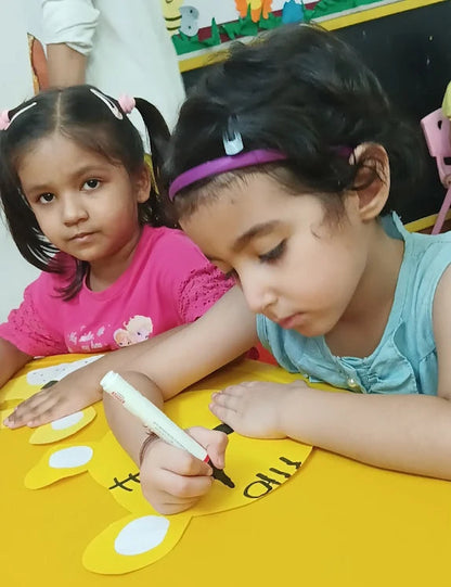 Two young girls sitting at a table with educational materials, one writing on a yellow surface.