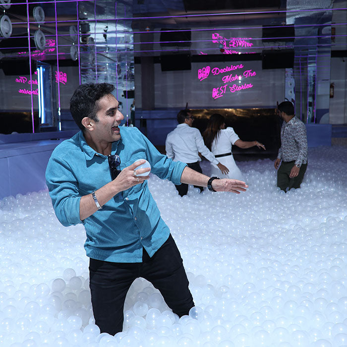 A man in a blue shirt throws a ball in a ball pit while others stand in the background.
