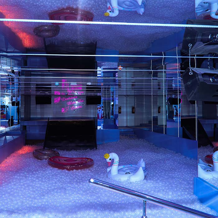 A ball pit filled with white balls, featuring swan inflatables and neon signs, with reflections from a mirrored ceiling.