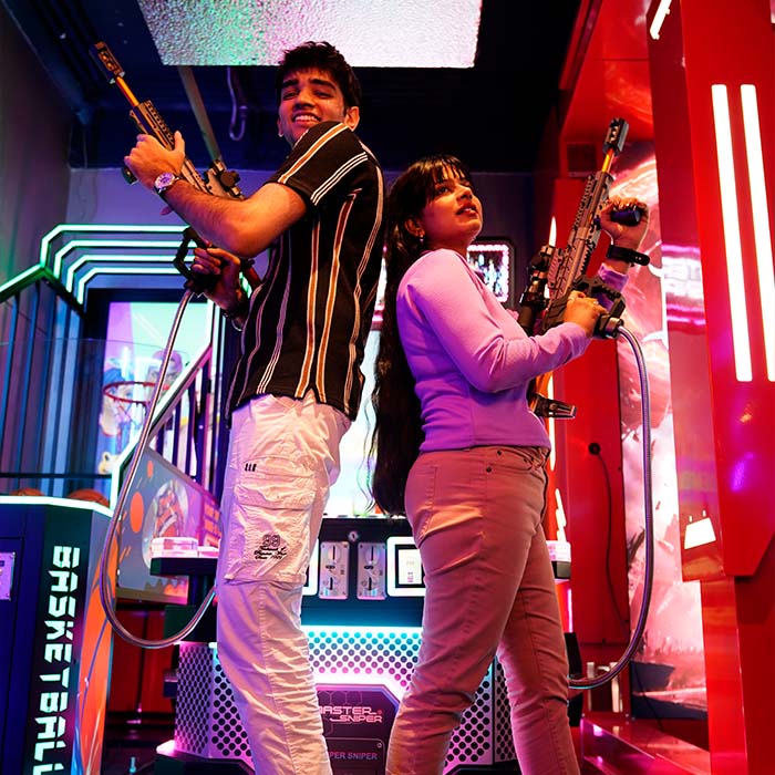 Two young adults play a shooting game in an arcade, holding toy guns.