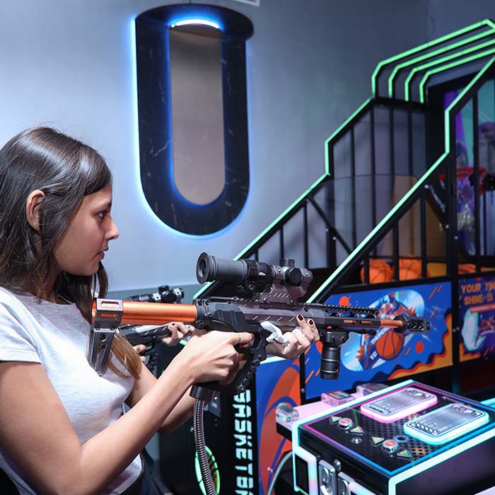 A young woman plays an arcade game, aiming a rifle at a screen. The game features neon lights and basketball imagery.