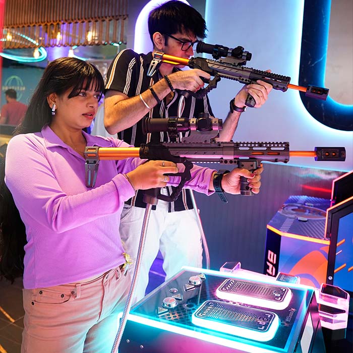 Two young adults play a shooting game at an arcade, holding toy rifles.