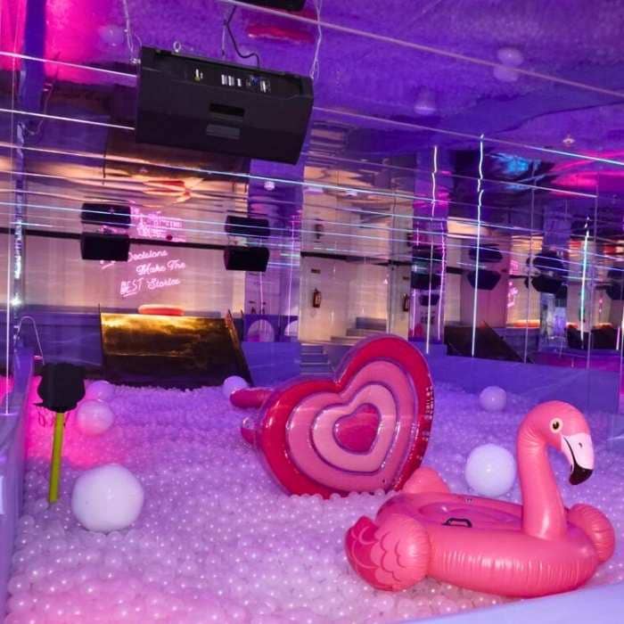 A ball pit filled with white balls, featuring a pink flamingo float and a heart-shaped float, under purple lighting.