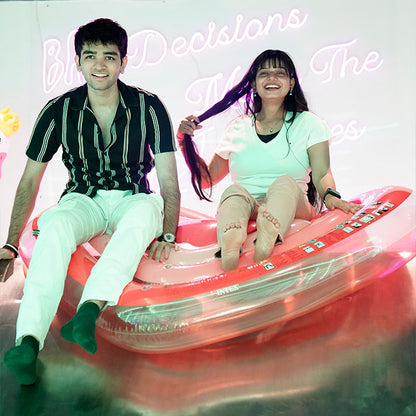 A young couple sits on a pink inflatable raft, with neon signs in the background.