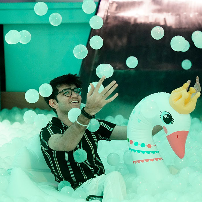 A young man in a ball pit throws translucent balls in the air next to a swan float.