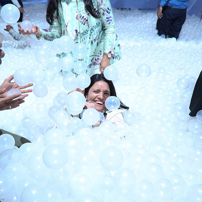 A woman laughs joyfully while submerged in a ball pit filled with white balls.