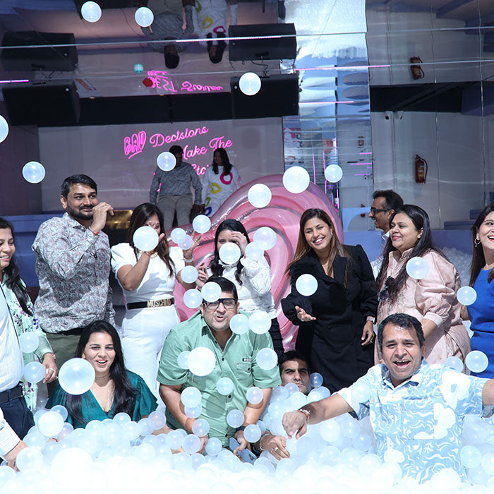 Adults in a ball pit, some holding bubbles, with a neon sign in the background.