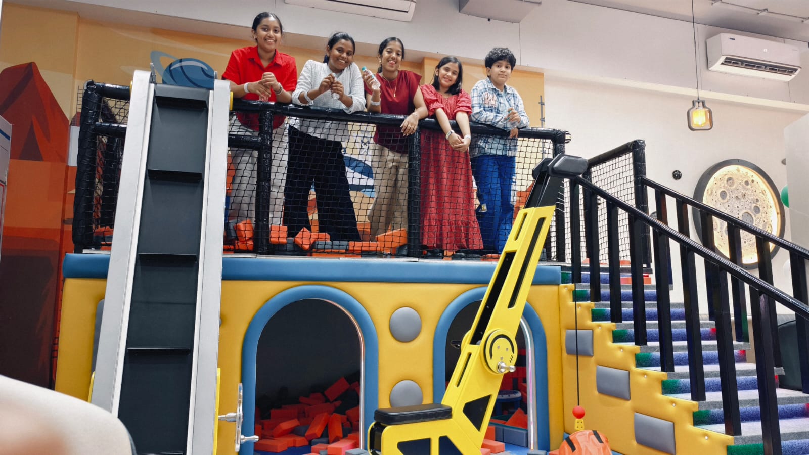 Children stand on a colorful indoor playground structure with a slide and stairs.