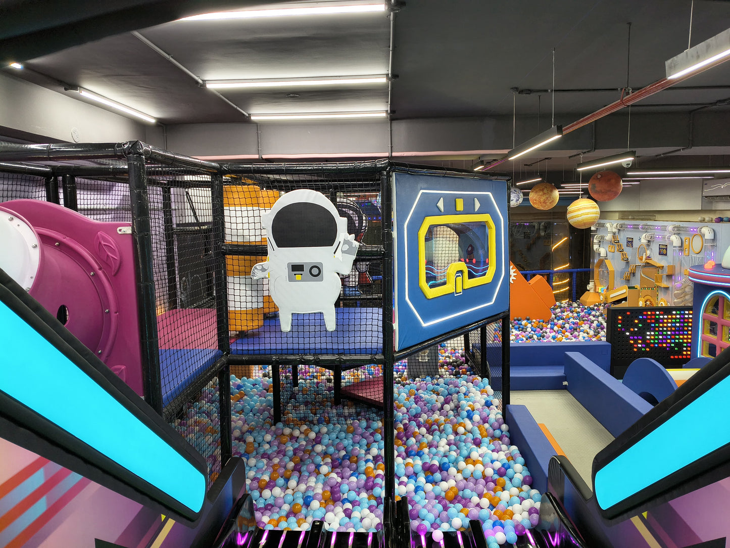 An indoor playground with a ball pit, slides, and space-themed decorations. Children can be seen playing in the ball pit.