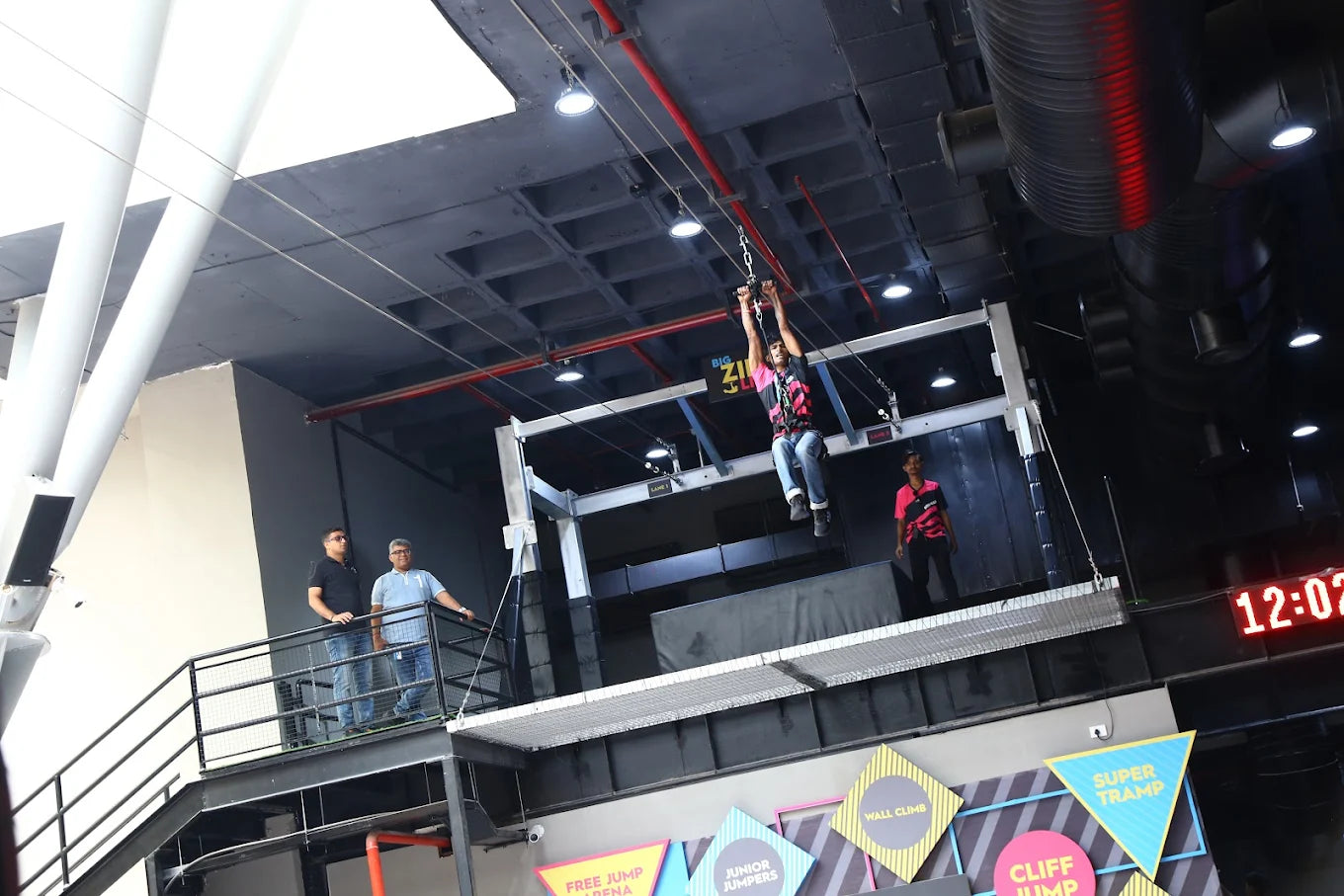 A young man ziplines across an indoor adventure park while others watch from a balcony.
