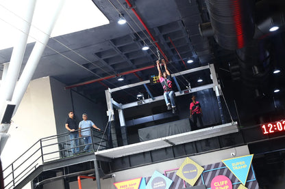 A young man ziplines across an indoor adventure park while others watch from a balcony.