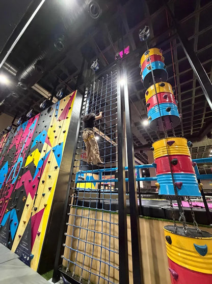 A person climbs a rope net next to a colorful climbing wall in an indoor adventure park.