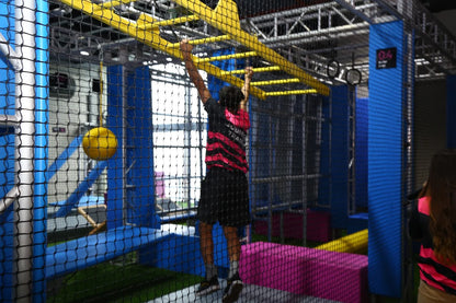 A young person climbs an obstacle course with yellow monkey bars and blue netting.