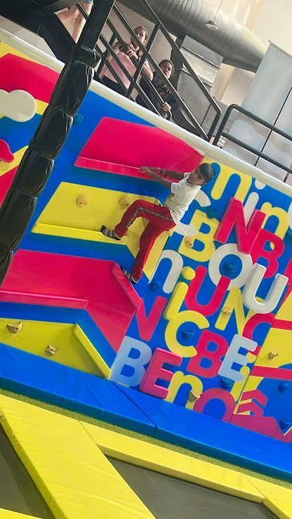 A young boy climbs a colorful indoor climbing wall while other children watch from above.