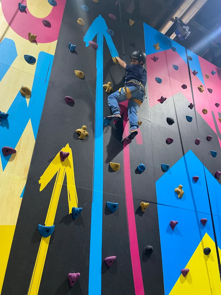 A child in a helmet and harness climbs a colorful indoor rock-climbing wall.