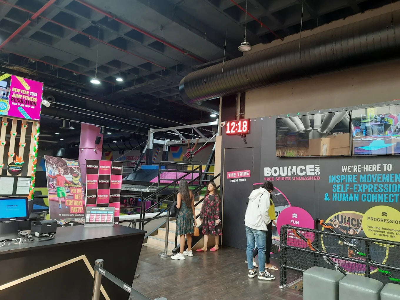 People at a trampoline park with a digital clock showing 12:18.