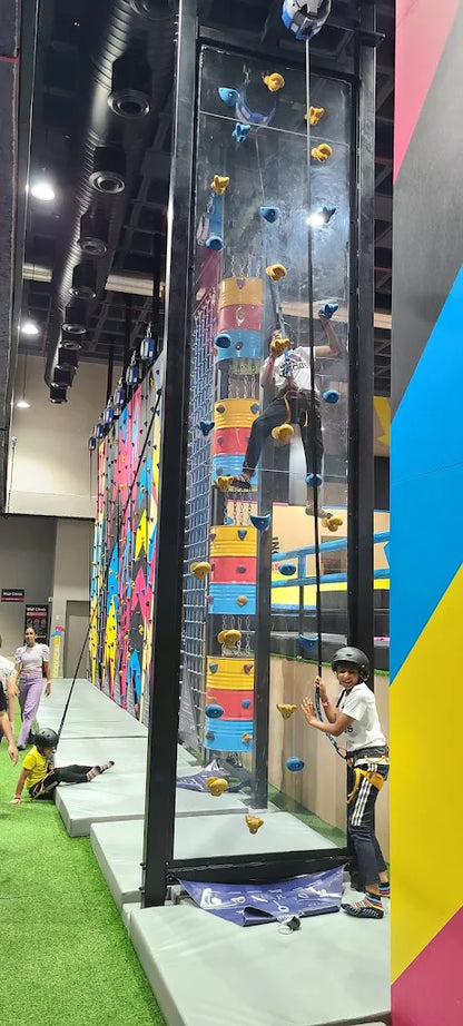 Children climb a colorful indoor rock wall with safety harnesses.