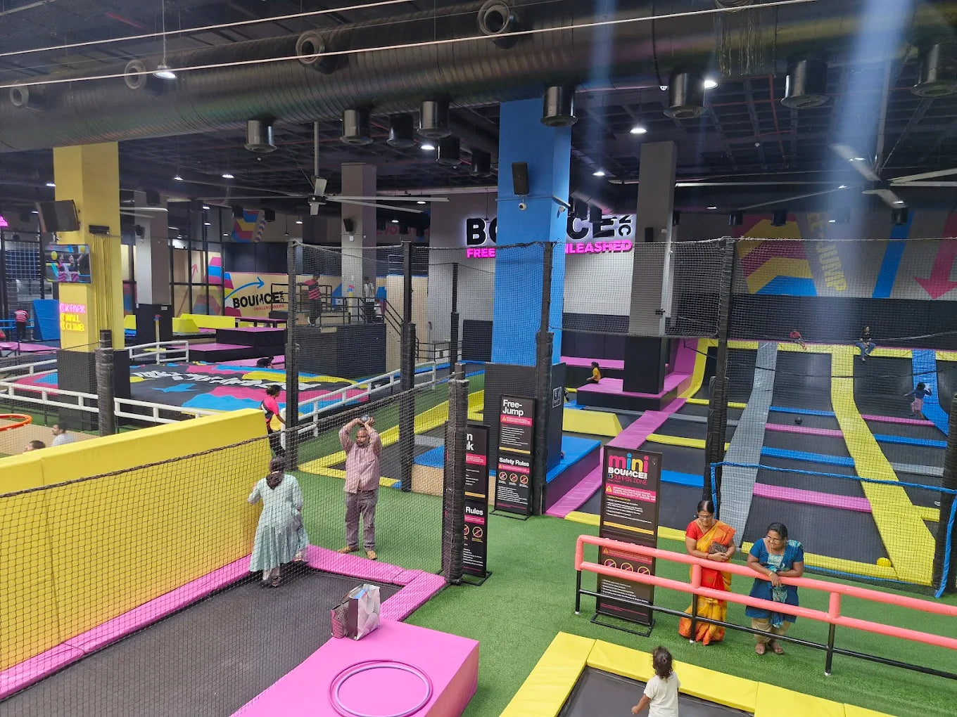 People of various ages enjoy a trampoline park with colorful bounce areas and safety nets.
