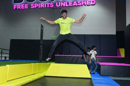 A young man jumps mid-air on a trampoline, with other young people in the background at an indoor trampoline park.